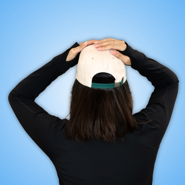 Vivian viewed from behind wearing a light beige baseball cap backwards with a dark green underbrim, against a solid light blue background.