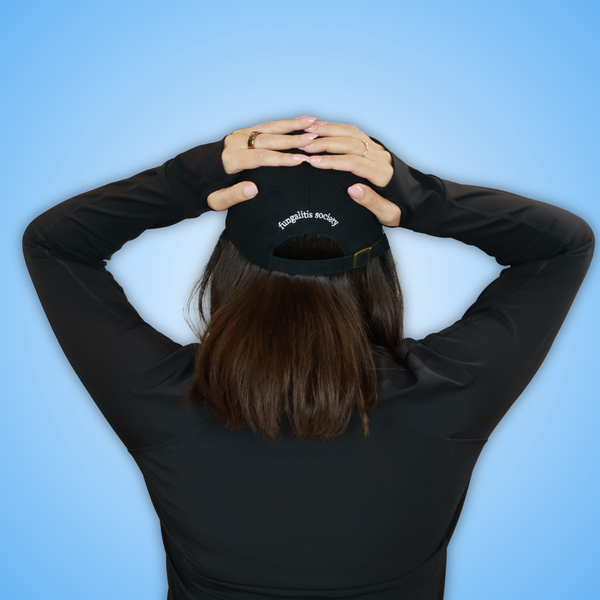 Vivian viewed from behind wearing a black long-sleeve top and a black baseball cap with 'fungalitis society' embroidered in white on the back, set against a solid light blue background.