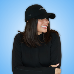 Vivian wearing a black baseball cap with white embroidered 'VC and 'vivchews' text, paired with a black long-sleeve top, set against a solid blue background.