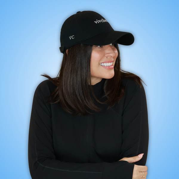 Vivian wearing a black baseball cap with white embroidered 'VC and 'vivchews' text, paired with a black long-sleeve top, set against a solid blue background.