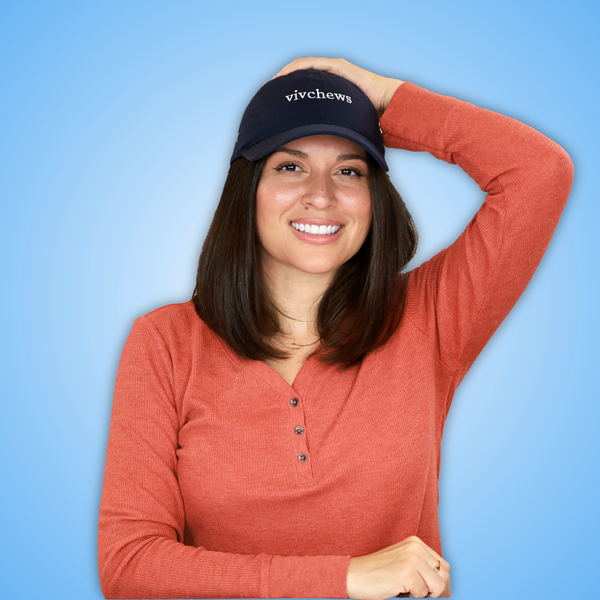 A smiling Vivian with shoulder-length brown hair wears a navy blue baseball cap with the white embroidered text 'vivchews' and a rust-orange ribbed henley shirt, posing against a light blue gradient background.