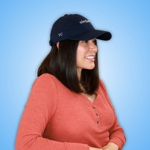 Vivian wearing a navy blue baseball cap with white embroidered text and a coral pink henley shirt, shown from the chest up against a solid light blue background.