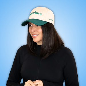 Vivian wearing a cream and green baseball cap with embroidered text, paired with a black long-sleeve top, set against a solid blue gradient background.