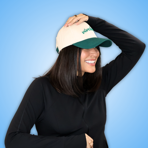 Vivian wearing a beige and green baseball cap with green embroidered text, paired with a black long-sleeve top, posing against a solid blue gradient background.